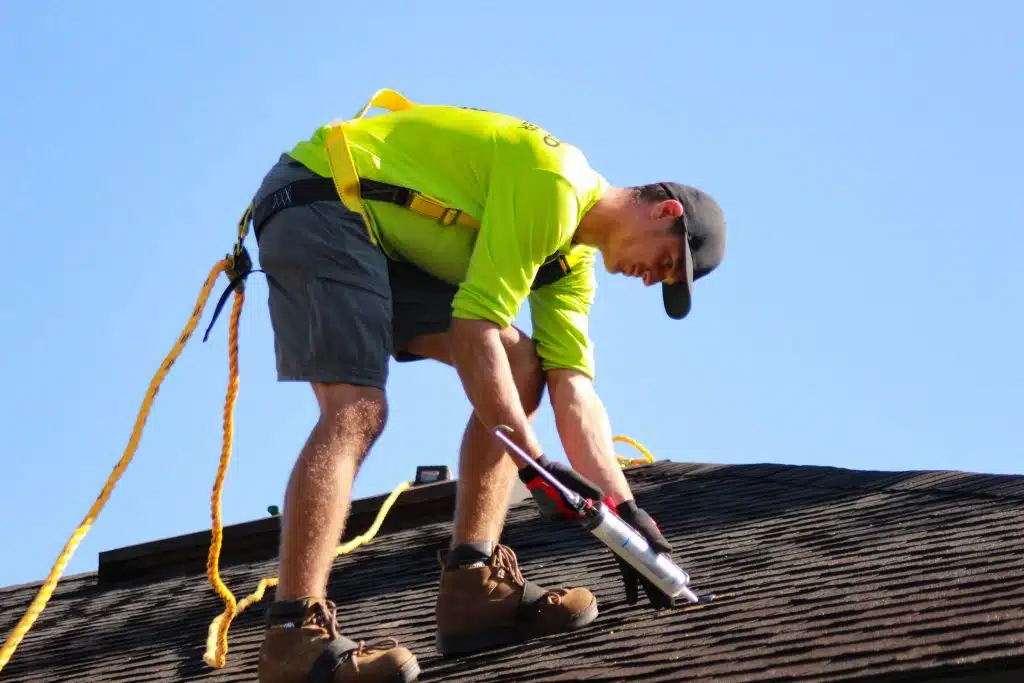 An architectural roofing contractor worker wearing a safety harness and high-visibility shirt applies sealant to a dark shingled roof with a caulking gun.