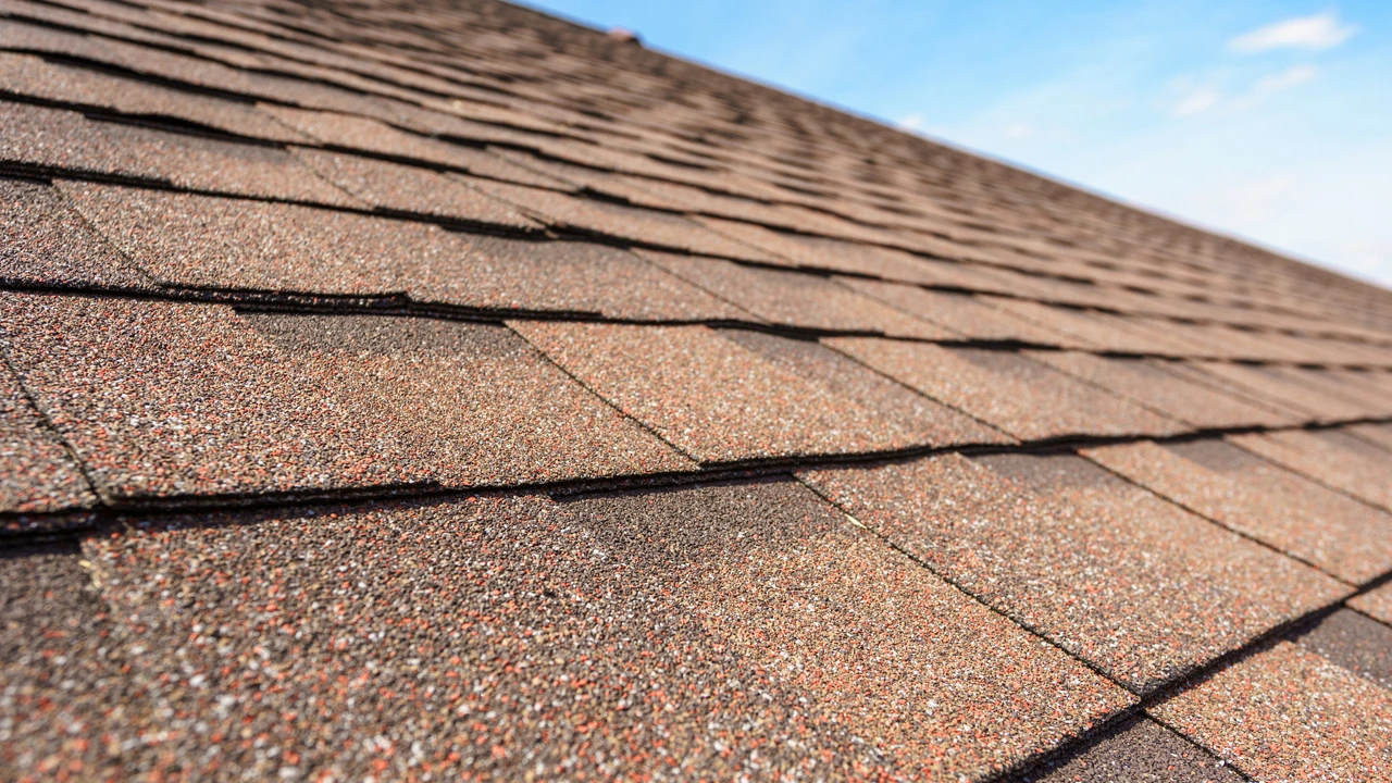 A close-up view of rows of brown asphalt shingles on a sloped roof under a clear blue sky.