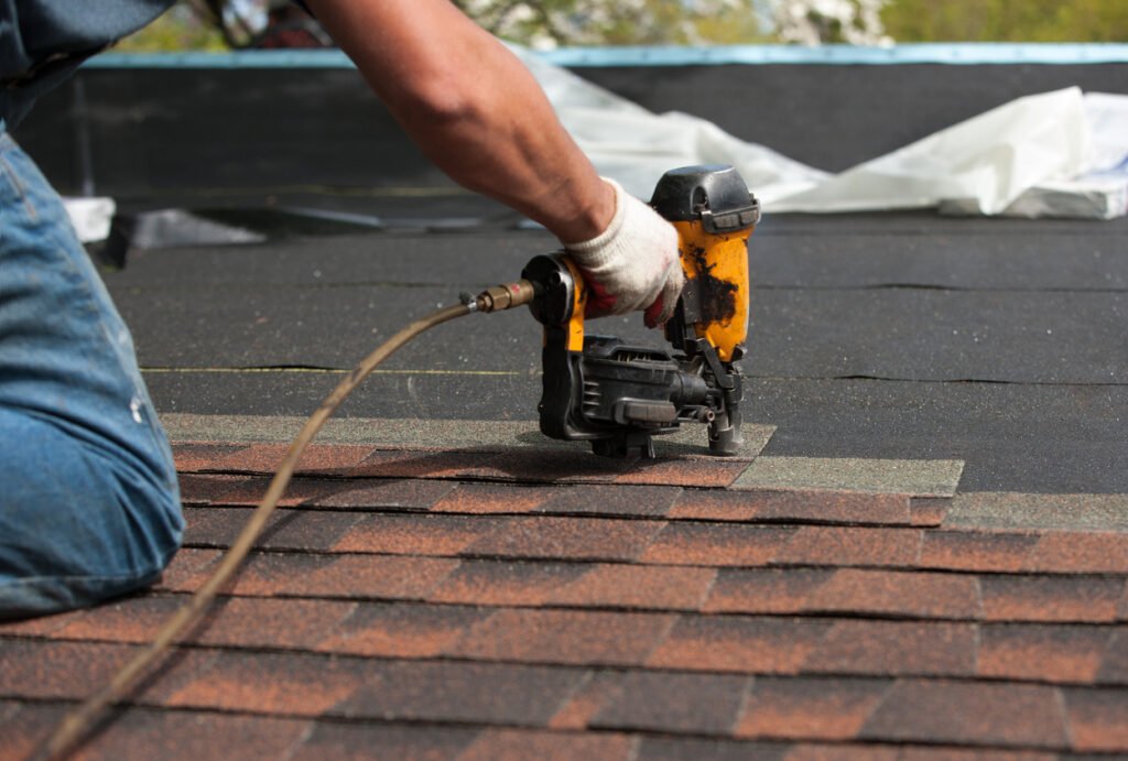 A roofer using a pneumatic nailer to secure reddish-brown asphalt shingles over black underlayment.