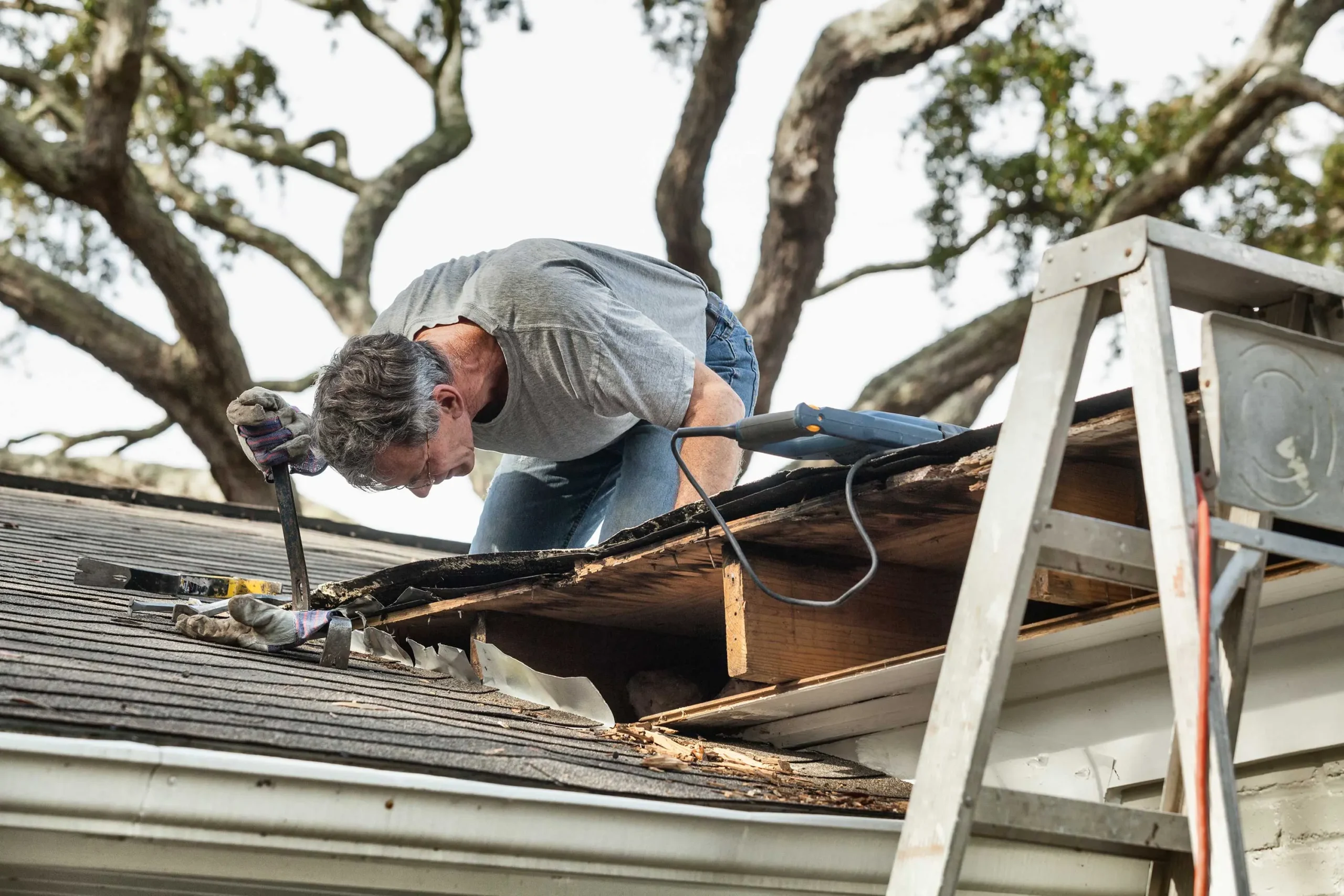 A worker on a roof uses a pry bar to remove damaged shingles and reveal the wooden structure underneath during a repair project.