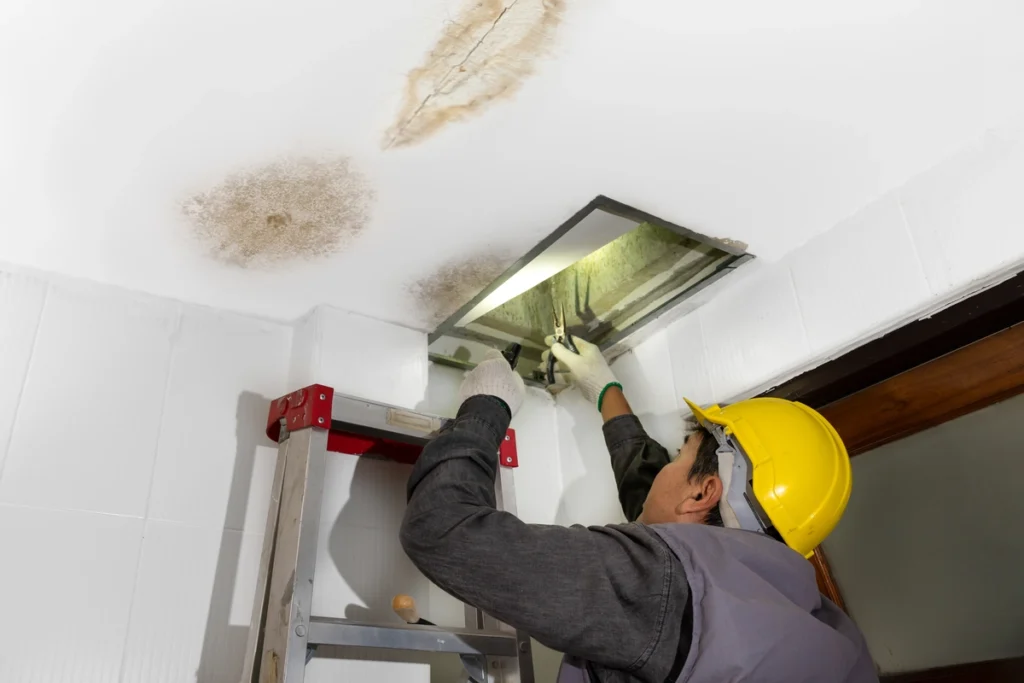 A worker on a ladder reaches into a ceiling access panel to inspect water damage and brown stains on the white ceiling.